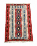Tpete Kilim -  Manufatura em lã, sobre trama de algodão, apresentando composição geométrica de inspiração tribal. Campo central em vermelho-terra, cinza e marfim, ornamentado por motivos escalonados em azul e marrom, emoldurados por faixas laterais com elementos losangulares repetidos. Acabamento com franjas nas extremidades. Medindo 1,80 x 1,24 m.