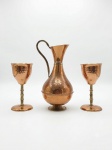 Conjunto em cobre maciço, composto por três peças, sendo uma ânfora/jarra central e dois cálices, um deles. Corpo de linhas clássicas com acabamento polido em cobre e bases torneadas. Jarra medindo 23 x 11 e Cálices medindo 15 x 7
