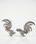 Par de esculturas de galos ornamentais. Em metal fundido com acabamento prateado envelhecido, representando galos em postura de exibição, com caudas longas e ricamente detalhadas em relevo, evidenciando o trabalho nas penas. As aves encontram-se sobre bases integradas de formato orgânico, compondo um conjunto decorativo de grande presença visual, ideal para aparadores, mesas ou estantes. Menor medindo 22 x 17 x 26 e Maior 25 x 18 x 18