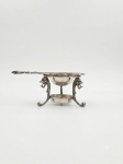 Coador de chá em metal prateado. Conjunto composto por suporte tripé com aro superior ornamentado em relevo floral, recipiente inferior e coador de chá em metal prateado com cabo ricamente decorado e concha perfurada para retenção das folhas de chá durante o serviço. Medindo 6 x 15 centímetros