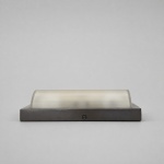 Medidas: 8 x 31,5 x 13,5 cm.Luminária de parede com estrutura retangular em metal de acabamento escurecido e difusor semicilíndrico em vidro fosco. A composição apresenta linhas horizontais depuradas, proporções alongadas e sistema de fixação integrado à face frontal. O vidro curvo proporciona difusão luminosa uniforme, enquanto o contraste entre a superfície translúcida e a estrutura metálica confere equilíbrio formal e presença arquitetônica à peça.