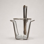 Medidas do balde: 15 cm de altura x 14 cm de diâmetro. Comprimento do pegador: 21 cm.Balde para gelo em vidro transparente de paredes espessas e base reforçada, acompanhado de suporte estrutural e pegador em metal. O suporte apresenta desenho geométrico aberto, com hastes verticais, aro superior e alça elevada para transporte. O pegador possui formato alongado e sistema de encaixe por argolas junto à estrutura. A combinação entre a transparência do vidro e o acabamento metálico confere ao conjunto equilíbrio de proporções, funcionalidade e presença visual.