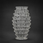 Medidas: 20x11Vaso escultórico em cristal transparente espesso, de formato vertical e perfil escalonado, estruturado por sucessivas faixas horizontais em relevo. A superfície apresenta modelagem orgânica, com saliências irregulares e bordas recortadas que intensificam os efeitos de refração, brilho e profundidade. Possui boca circular, corpo de proporções robustas e base anelar, reunindo transparência acentuada, textura expressiva e composição arquitetônica de forte presença visual.