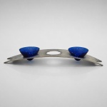 Conjunto de servir composto por suporte arqueado em metal e duas taças removíveis em vidro azul-cobalto, assentadas sobre hastes cilíndricas em vidro transparente. Essa combinação de recipientes coloridos encaixados em armação metálica integra a tradição dos conjuntos de condimentos e aperitivos destinados ao serviço de mesa. A peça mede 7 cm de altura x 48 cm de comprimento x 14 cm de profundidade. O suporte apresenta estrutura horizontal alongada, três aberturas circulares e extremidades recortadas, enquanto o contraste entre o azul intenso, a transparência do vidro e o acabamento metálico confere equilíbrio formal e presença visual ao conjunto.