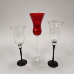 Trio de porta-velas decorativos em vidro, constituído por recipientes superiores de perfil tulipa sustentados por hastes longas e bases circulares. As peças medem 46 cm, 42 cm e 38 cm de altura, todas com 12 cm de diâmetro. O conjunto combina dois exemplares em vidro incolor com hastes e bases pretas e um exemplar central em vidro vermelho-rubi com haste e base transparentes. A alternância cromática, a gradação das alturas e as linhas alongadas formam uma composição equilibrada, concebida para receber velas votivas ou flutuantes e produzir efeito luminoso através dos bojos translúcidos. Essa configuração integra a categoria dos porta-velas elevados do tipo hurricane, reconhecidos pelos amplos recipientes de vidro sobre pedestal.