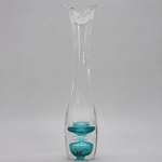 Vaso escultórico da manufatura sueca Vas Vitreum, executado artesanalmente em cristal incolor espesso, com composição vertical alongada e inclusões internas em tonalidade turquesa. A peça mede 34 cm de altura x 6 cm de diâmetro máximo. Apresenta base maciça, sucessão de elementos circulares suspensos no interior, colo estreito e prolongado e boca assimétrica modelada em curvas orgânicas. A transparência acentuada e a disposição das inclusões produzem efeitos de refração, profundidade e movimento ao longo do corpo. Preserva etiqueta original VAS VITREUM  Crystal Sweden  Vadstena, identificando a manufatura fundada em 1985 e reconhecida pela produção manual de cristal artístico na Suécia.
