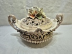 Peça decorativa italiana em faiança Bassano, modelo R/242, com corpo rendado em estilo barroco, alças ornamentadas e tampa vazada com flores modeladas à mão, apresentando craquelês típicos da idade e pequenos desgastes compatíveis com o uso, ideal para decoração clássica ou coleção Medindo 21cm x 14cm de altura. Com pequenos bicados.