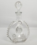 BACCARAT  - COM MARCAS DAS MANUFATURA DECANTADOR DE CONHAQUE LOUIS XIII EM CRISTAL BACCARAT, ENCOMENDA PARA REMY MARTIN, COM FLOR DE LIS EM RELEVO E LATERAIS COM ARREMATES EM SERRILHA NAS LATERAIS. ROLHA EM FORMA DE LÁGRIMA. MARCA ACIDADA NA BASE.  FRANÇA, SEC. XX. 28 CM DE ALTURA. ATENTE QUE EXISTE UM DISCRETO BICADO EM UM DOS OITO MEDALHÕES  COM FLORES DE LIZ DISPOSTOS NA GARRAFA. NOTA: O design do decanter Baccarat encomendado pela Remy Martin não nasceu da imaginação moderna. Ele é a réplica exata de uma garrafa metálica de carapaça (um frasco de metal) perdida no ano de 1569 durante a Batalha de Jarnac, na França. Em 1850, o fundador do conhaque, Paul-Émile Rémy Martin, comprou esse frasco histórico de um camponês que o encontrou enterrado em um campo. Paul-Émile usou essa silhueta única para engarrafar sua bebida mais prestigiada a partir de 1874. A fabricação de cada garrafa padrão exige uma coreografia perfeita. São necessários exatamente 11 artesãos trabalhando em total sincronia para soprar, moldar e cortar o cristal manualmente enquanto ele ainda está incandescente. Devido a esse processo estritamente manual, não existem dois decanters perfeitamente iguais no mundo. Cada decanter produzido pela Baccarat recebe uma numeração exclusiva gravada na base, que coincide exatamente com o número gravado em sua respectiva tampa de cristal. Essa numeração serve como certificado de autenticidade para colecionadores em leilões pelo mundo.