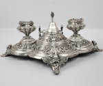 GRANDE TINTEIRO EM  PRATA DE LEI COM MARCAS DE CONTRASTE DA ALEMANHA SEGUNDA METADE DO SEC. XIX. DECORAÇÃO COM FIGURAS DE HOMENS VERDES DE CUJA BOCA EMERGEM FRUTOS DE CARVALHO E DE LOURO. A TODA VOLTA ROCAILLES RELEVADAS. DOTADO DE DOIS FRASCOS COM FEITIO DE ANFORAS MEDICI CONTENDO EM SEU INTERIOR RECIPIENTES EM VIDRO ARTISTICO , AO CENTRO UMA SINETA E AO FUNDO PORTA PENAS. ALEMANHA, SEC. XIX. 37 CM DE COMPRIMENTO. 875 GNOTA: NOTA: Homem Verde é uma escultura, desenho, ou outra representação de um rosto rodeado por folhas. Ramos ou cipós podem brotar pelo nariz, boca, narinas ou outras partes do rosto e estes brotos podem conter flores ou frutas. Comumente usados como ornamentos decorativos, Homens Verdes são frequentemente encontrados em esculturas, igrejas e outros edifícios (tanto seculares quanto eclesiásticos). "O Homem Verde" também é um nome popular para pubs ingleses e diferentes interpretações do nome aparecem em letreiros de pousadas, que, algumas vezes, mostram uma figura inteira ao invés de somente a cabeça.O motivo Homem Verde tem muitas variações. Encontrado em muitas culturas ao redor do mundo, o Homem Verde é muitas vezes relacionado a divindades de natureza vegetal em diferentes culturas ao longo dos tempos. Essencialmente, ele é interpretado como um símbolo de renascimento, representando o ciclo de crescimento a cada primavera. Especula-se que a mitologia do Homem Verde desenvolveu-se independentemente nas tradições de culturas ancestrais separadas e evoluiu para a grande variedade de exemplos encontrados ao longo da história.