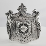 PALÁCIO DE SÃO CRISTÓVÃO  SALA DO TRONO REFORMULADA NA DECADA DE 1840 - PRATA DE LEI INGLESA. PRENDEDOR DE REPOSTEIRO EM PRATA DE LEI COM FEITIO DE BRASÃO IMPERIAL OSTENTANDO 19 ESTRELAS QUE REPRESENTAVAM AS PROVINCIAS DO IMPERIO DO BRASIL APLICADO SOBRE MANTO DE ARMINHO ACOMODADO SOB PÁLIO COM FESTÕES E ENCIMADO PELA COROA IMPERIAL QUE SEGUE AINDA O MODELO DA COROA DE DOM PEDRO I. GUARNECEU A SALA DO TRONO DO PALÁCIO DE SÃO CRISTÓVÃO. MARCAS DE CONTRASTE PARA CIDADE DE SHEFFIELD LETRA DATA PARA O ANO DE 1840. PRATEIRO THOMAS CRESWICK. MARCAS DO PRATEIRO NA PARTE POSTERUIR E TAMBÉM NA FRONTAL. AINDA UMA INSCRIÇ;AO ATRÁS: "END N. 2". DÉCADA DE 1840 MARCOU UMA INTENSA CAMPANHA PARA ENOBRECER O DECADENTE PALÁCIO DE SÃO CRISTÓVÃO QUE DESDE A ÉPOCA DA IMPERATRIZ LEOPOLDINA NÃO FAZIA NOVAS AQUISIÇÕES. O MOTIVO DESTA REVITALIZAÇÃO FOI O GOLPE DA MAIORIDADE QUE LEVOU DOM PEDRO II A SER COROADO AOS 14 ANOS EM 18 DE JULHO DE 1841E E SEU CASAMENTO COM A IMPERATRIZ TEREZA CHRISTINA NO RIO DE JANEIRO EM 4 DE SETEMBRO DE 1843. ERA NECESSÁRIO TRAZER LUSTRO A CASA REINANTE IMPERIAL. O PALÁCIO ROTO E DESPROVIDO DE ALFAIAS ERA UM SÍMOBOLO DE UM REGIME DECADENTE QUE JAMAIS PODERIA SER OSTENTADO NO PRIMEIRO ANO DE REINADO DO PRIMEIRO IMPERADOR BRASILEIRO NASCIDO EM NOSSAS TERRAS. PARA SAUDAR O NOVO IMPERADOR E IMPERATRIZ , O ANTIGO PAÇO DE SÃO CRISTÓVÃO E A CIDADE DO RIO DE JANEIRO PASSARAM POR REMODELAÇÕES. O PALÁCIO FOI PREPARADO PARA O GRANDE BAILE DE GALA, ENQUANTO A CAPITAL ADOTOU A ESTÉTICA NEOCLÁSSICA PARA CONSOLIDAR O PODER DO NOVO IMPÉRIO. UMA CARRUAGEM  DE GALA COM MUITOS ADORNOS EM PRATA FOI ENOCMENDADA NA CIDADE DE LONDRES. EM SHEFFIELD OS PRATEIROS THOMAS, JAMES & NATHANIEL CRESWICK PRODUZIRAM PEÇAS DE APARATO E ALFAIAS PARA MESA EM PRATA DE LEI. AS REFORMAS ESTRUTURAIS E E A ENCOMENDA DA PRATARIA INGLESA REPRESENETAM UMA DAS FACES MAIS IMPORTANTES DA MODEERNIZAÇÃO DO PÃO DE SÃO CRISTÓVÃO. UM MANUSCRITO DA ÉPOCA QUE PERNTENCE AO ARQUIVO NACIONAL DESTACA ESSAS REFORMAS: ABRIO COMODOS, UNIO MAIS APOSENTOS, E COPAS NESTA FORMA PARA SUBSTITUIR AS ANTIGAS E FEZ UM ARRECADAMENTO RICO. COPAS COM SERVIÇO DE PRATA MACIÇA QUE O DITO O MORDOMO MANDARA FAZER NA INGLATERRA Q' SERVE DO ACTUAL IMPERADOR, E TANTES AS MAIS Q' MUITAS DELAS TEM COMPRADO CUJO ... RENDE A 60.O KG. BARBOZA HAVIA DOTADO A CASA MPERIAL COM BOTÕES DE OURO MESMOS ... DE GLAA . E FEZ SOB SUA ADMINISTRAÇÃO QUE TIVESSEMOS 12 CARRUAGENS TIRADAS A 8 E A 6 CAVALLOS D' RAÇA. ENTRETANTO O MESMO MANUSCRITO REFERE-SE A PESSOA DO IMPERADOR QUE DESCENDE DE DUAS CASAS OLHÃO PARA RAÇA E NÃO PARA OPULENCIA. SOB A SUPERVISÃO O MORDOMO  PAULO BARBOSA, A FACHADA FRONTAL DO PALÁCIO COMEÇOU A SER REMODELADA E O PAVIMENTO SUPERIOR FOI ACRESCIDO, COM A CONTRATAÇÃO DE ARTISTAS E ARQUITETOS COMO MANUEL DE ARAÚJO PORTO ALEGRE (BARÃO DE SANTO ANGELO). A GRANDE TRANSFORMAÇÃO ARTÍSTICA DA SALA DO TRONO FICOU A CARGO DO CENÓGRAFO E PINTOR ITALIANO MARIO BRAGALDI, QUE UTILIZOU A TÉCNICA DE TROMPE-L'IL (ILUSÃO DE ÓTICA QUE SIMULA ALTO-RELEVO EM SUPERFÍCIES PLANAS). TETO E PAREDES: FORAM DECORADOS EM ESTILO RENASCENTISTA COM ALEGORIAS MITOLÓGICAS QUE INCLUÍAM DIVINDADES COMO JÚPITER, VÊNUS, CUPIDO, MERCÚRIO E MINERVA. QUATRO ÂNGULOS DO TETO, FORAM PINTADAS FIGURAS REPRESENTANDO A JUSTIÇA, FORTALEZA, TEMPERANÇA E PRUDÊNCIA, SERVINDO COMO UM LEMBRETE VISUAL DAS QUALIDADES QUE DEVERIAM GUIAR O GOVERNO DO JOVEM MONARCA. O MOBILIÁRIO FOI PLANEJADO PARA REFLETIR A NOVA IDENTIDADE DO IMPÉRIO. O TRONO DE D. PEDRO II ERA UMA OBRA-PRIMA DE TALHA DOURADA, COM ENCOSTO, ASSENTO E BRAÇOS INTEIRAMENTE REVESTIDOS DE VELUDO VERDE (A COR DA CASA DE BRAGANÇA E DO BRASÃO IMPERIAL). O ENCOSTO, EM FORMATO OVAL, EXIBIA NO CENTRO O MONOGRAMA BORDADO P II. O  PLANEJAMENTO DA MORDOMIA VISAVA DAR VAZÃO AO ENORME FLUXO DE VISITANTES. A SALA DO TRONO CONECTAVA-SE ESTRATEGICAMENTE À SALA DOS EMBAIXADORES (CORPO DIPLOMÁTICO) PARA ORGANIZAR O PROTOCOLO DE CUMPRIMENTO AO IMPERADOR. AS COMEMORAÇÕES DA COROAÇÃO ESTENDERAM-SE POR NOVE DIAS E CULMINARAM EM 24 DE JULHO DE 1841 COM UM MONUMENTAL BAILE DE GALA NO PALÁCIO, ONDE A NOVA ORNAMENTAÇÃO DA SALA DO TRONO SERVIU DE CENÁRIO PARA A CONSOLIDAÇÃO SIMBÓLICA DO PODER DE D. PEDRO II. O TRONO ERA O PONTO CENTRAL E FICAVA POSICIONADO NO FUNDO DA SALA, SOBRE UM TABLADO ELEVADO (ESTRADO) COM DEGRAUS. ACIMA DO TRONO, UMA GRANDE ESTRUTURA DE MADEIRA DOURADA SUSTENTAVA UM MAJESTOSO CORTINADO DE VELUDO VERDE E ARMINHO QUE DESCIA PELAS LATERAIS, EMOLDURANDO O ASSENTO DO MONARCA. A SALA POSSUÍA VÁRIAS PORTAS DE COMUNICAÇÃO QUE FACILITAVAM O RÍGIDO PROTOCOLO DA CORTE. ELA CONECTAVA-SE DIRETAMENTE À SALA DOS EMBAIXADORES (ONDE O CORPO DIPLOMÁTICO AGUARDAVA) E A OUTRAS SALAS DE APARATO, PERMITINDO QUE A FILA PARA O CUMPRIMENTO AO IMPERADOR FLUÍSSE DE MANEIRA ORGANIZADA, SEM TUMULTOS. AS GROSSAS CORTINAS DE VELUDO EMOLDURAVAM AS JANELAS AMARRADAS POR GANCHOS EM PRATA DE LEI  ATADOS A FESTÕES. ERAM DECORADOS COM AS ARMAS DO IMPÉRIO CONFECCIONADAS PELOS PRATEIROS DA CORTE APLICADAS SOBRE ESCUDOS ENCOMENDADOS A PRATEIROS DA INGLATERRA. DECADA DE 1840. 10 X 7,5 CM 75 GNOTA: Festejos da Coroação de Dom Pedro II no Paço Imperial em 1841. Dom Pedro II, embora jovem, estava profundamente commovido e entristecido, assistia a todas convulsoes politicas do Brasil Regencial, pensando nos meios de lhes por um termo. Esforcos intellectuaes de tal natureza desenvolveram cedo suas faculdades excepcionaes e fizeram delle um homem antes de tempo. Inspirou. assim. inteira confianca ao Parlamento, que. temeroso de uma parte do paiz sempre em agitacao. julgou necessário encurtar de tres annos a epoca legal para a maioridade do jovem imperador. D.Pedro II nao passava dos quinze annos em 1840. A guerra acabrunhava uma das províncias mais importantes do Imperio, o Rio Grande do Sul, propagando-se pelas demais. Uma angustia geral opprimia a nacao. Todo o progresso estava entravado. Movido de patriotismo e sentindo-se alias, a altura da missao para que era solicitado tao prematuramente, acceitoua D, Pedro II, corajosamente, a hora do perigo. Reunidas as duas Camaras em assemblea geral, aos 23 de julho de 1840 foi declarada a maioridade, organizando elle o seu primeiro ministerio.A 18 de julho do anno seguinte. realizou-se. na Cathedral do Rio, a cerimonia da sagracao e coroacao de D. Pedro II. no meio do enthusiasmo geral. As festas se prolongaram por varios dias. Trecho do livro Livro: Vida de Dom Pedro II - Benjamin Mosse, 1892