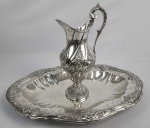 ELEGANTE GOMIL E LAVANDA DE APARATO  EM PRATA DE LEI  COM MARCAS DE CONTRASTE  ESTILO PARA A CIDADE DO RIO DE JANEIRO COM INICIAIS DO PRATEIRO F.I SOB COROA IMPERIAL DE DOM PEDRO I REFERENCIADAS POR MOITINHO COMO BR226 DATAVEL A PARTIR DE 1830. PEÇAS MAGNIFICAS COM GRANDE QUALIDADE NA EXECUÇÃO. GOMIL DE CORPO ESPIRALADO DECORADO COM ALETAS, ENROLAMENTOS VEGETALISTAS, GUIRALANDA DE LAUREU CAPRICHOSAMENTE CINZELADA. ELEGANTE ALÇA EM CARAPETA.  LAVANDA DE FEITIOO OBLONGO COM BORDO RECORTADO E DECORADO COM CONCHEADOS E FLORES. TAMBEM ENROAMENTOS VEGETALISTAS. UMA PEÇA DE SALÃO PARA USO DE ABLOUÇAO DAS MÃOS AO JANTAR. BRASIL, DEC. 1830. 45 CM DE COMPRIMENTO (LAVANDA), 1970 G.NOTA: Antes da existência da água encanada e dos banheiros modernos, o gomil acompanhado de sua bacia desempenhava papel central na rotina da nobreza e aristocracia. Utilizado no ritual do lavabo, o gomil servia para verter água (frequentemente perfumada com pétalas ou essências) sobre as mãos dos comensais antes e depois das refeições em banquetes de gala, bem como nos aposentos privados para a higiene diária. Mais do que utilitário, era um símbolo de ostentação, civilidade e status social. . Antes de servirem o banquete, os criados circulavam entre os convidados segurando o gomil com elegância para derramar suavemente um fio de água perfumada sobre as mãos dos comensais vertendo-a nesta  imponente bacia. Ter uma peça dessas reluzindo no salão não era apenas uma questão de higiene, mas a declaração máxima de poder, bom gosto e etiqueta de uma época. Dificilmente o gomil era preenchido com água comum. Usava-se água morna infusionada com pétalas de rosa, flor de laranjeira, cravo ou alecrim, criando uma experiência sensorial de higiene e perfume para os convidados. Nas grandes cortes europeias e na Imperial Quinta da Boa Vista no Brasil, segurar o gomil e a bacia para o monarca não era tarefa de um criado comum, mas sim um privilégio concedido a nobres de alta linhagem (como o Camareiro-Mór ou o Trinchante). O gomil é um dos raros objetos que transitam com a mesma elegância no ambiente profano (salões aristocráticos) e no sagrado (utilizado pelo sacerdote no ritual do Lavabo durante a celebração da Missa católica).