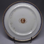 SERVIÇO DE DOM JOÃO VI - VISTA PEQUENA - PRATO EM PORCELANA CIA DAS ÍNDIAS QUE PERTENCEU AO SERVIÇO REAL DE D. JOÃO VI, CONHECIDO COMO VISTA PEQUENA. PERÍODO, QING, JIAQING (1796-1820). ABA COM BARRADO GEOMÉTRICO EM AZUL REALÇADO EM OURO, SUCEDIDO NA BORDA POR FRISO EM OURO. EM DIREÇÃO A CALDEIRA NOVO BARRADO FORMADO POR ELOS EM OURO ENTREMEADOS POR LOSANGO PONTILHADO EM AZUL. NA CALDEIRA DECORAÇÃO EM ELIPSE CONSTRUÍDA EM OURO E EM RESERVA, CENA NA TONALIDADE ROUGE DE FEUR COM CASA E ÁRVORES. REPRODUZIDO NO LIVRO LOUÇA DA ARISTOCRACIA DO BRASIL POR JENNY DREYFUS. CHINA, SEC. XIX, 25 CM DE DIAMETRO NOTA: De acordo com Leila Mezan Algranti, em seu artigo: Em torno da mesa do rei: artefatos, convivialidade e celebração no Rio de Janeiro joanino, em 26 de abril de 1821, após doze anos de estadia no Rio de Janeiro, iniciava-se a viagem de regresso de D. João VI a Portugal a fim de, mais uma vez, preservar sua Coroa e domínios. Para a autora, a partida da Corte foi precedida de muitas indecisões políticas e de intensos preparativos de ordem prática, da mesma forma como acontecera ao viajar para o Brasil, em 1807. Naquela ocasião, em meio aos riscos de invasão do Exército napoleônico, foi necessário acomodar nas embarcações não só os ilustres passageiros, mas também um conjunto imenso e variado de coisas destinadas ao uso particular de seus proprietários, bem como da Casa Real.Um conjunto de peças de prata, roupas de mesa e artigos de copa, recém-chegados do Rio de Janeiro, viajou para Lisboa sob a incumbência de Joze de Britto, fiel da mantearia do rei, e foi entregue no pátio das cozinhas do Palácio da Ajuda a Joze Caetano Trigo, também funcionário do mesmo departamento da Casa Real, o qual realizou o registro das peças e o assinou por ordem de João Lourenço de Andrade que, por sua vez, recebeu todo o material em nome de Caetano Joze de Campos e Andrade Pinto, manteeiro da Casa Real, naquele momento. O título do registro Inventário da Prata, Roupa e mais trem pertencente à Mantearia de Sua Majestade que veio do Ryo de Janeiro indica claramente que, ao retornar do Rio de Janeiro, D. João VI fez embarcar objetos de prata, os quais constituíam parte ou a totalidade de um serviço de mesa. Entretando deixou no país quase todos os serviços de porcelana adquiridos para seu uso aqui ou trasladados de Portugal quando de sua fuga. O conjunto poderia conter elementos da famosa baixela Germain, assim denominada por ter sido encomendada a François Germain por D. José I, em 1756, após o terremoto ter destruído a baixela de D. João V, fabricada pelo ourives de Luís XIV (Thomas Germain) e pai do prateiro de D. José. A suposição de que poderia haver peças da baixela Germain de D. José I entre os artefatos registrados no inventário advém do fato de que há informações sobre seu transporte para o Brasil, bem como estudos referentes à divisão da mesma entre D. João VI e D. Pedro I, antes do retorno do monarca a Portugal. Os bens que permaneceram no Brasil, por sua vez, teriam sido gravados com as insígnias imperiais e o monograma P1º. De acordo com Leonor d'Orey, considerando-se o que atualmente se encontra preservado desta baixela em Portugal, o lote do imperador do Brasil era menor, embora incluísse várias peças muito prestigiosas, as quais se encontram dispersas em coleções particulares e de museus estrangeiros, vendidas após a deposição de D. Pedro II. Sabe-se igualmente que entre a prataria de mesa de grande aparato pertencente à Casa Real havia peças da baixela dos duques de Aveiro, confiscada pela Coroa, após o atentado a D. José em 1759,15 assim como peças denominadas avulsas. Por outro lado, as baixelas para D. Pedro I foram encomendadas às famosas casas francesas, embora a técnica dos ourives radicados no Brasil fosse excelente, como apontou Francisco Marques dos Santos. Segundo o mesmo autor, muitas dessas peças emigraram, mas algumas voltaram. Havia, portanto, na corte no Rio de Janeiro, mais de um serviço de mesa em prata, sendo que um conjunto significativo dessas peças atravessou o Atlântico pelo menos duas vezes, a exemplo do que se sucedeu com a Família Real. Além disso, vários desses objetos foram possivelmente fabricados na França e na Inglaterra, uma vez que os prateiros portugueses nem sempre eram considerados, na época, suficientemente habilidosos para agradar o sofisticado gosto da alta nobreza portuguesa, consumidora de produtos de luxo importados não só de outros países da Europa, como do Oriente. Embora, como apontou Gonçalo de Vasconcelos e Sousa, houvesse prateiros de renome em Portugal, fabricando artefatos de mesa e mesmo baixelas completas, cuja qualidade do trabalho atingiu durante o século das Luzes um dos seus momentos mais importantes. De qualquer modo, a mesa da Casa Real e aquelas de outros membros das elites europeias e luso-brasileiras eram há muito tempo aparamentadas com artefatos de prata estrangeiros, bem como com porcelanas e vidros chineses, germânicos e franceses. Como bem apontou Nuno Vassallo e Silva, em seu estudo sobre a ourivesaria da mantearia da Casa de Aveiro, não é possível refletir sobre as baixelas de prata ou de porcelana usadas na mesa real sem levar em conta a conexão dessa indústria com as manufaturas europeias e orientais. Assim, é possível dizer que os objetos de luxo destinados ao serviço de mesa da Corte joanina, no Rio de Janeiro, resultavam de um circuito comercial e de comunicação que interligava diferentes impérios coloniais (português, inglês, francês), os quais se estendiam por três continentes: Europa, Ásia e América. No caso dos artefatos de prata que nos interessam comentar neste estudo, é provável que a matéria-prima utilizada fosse originária da América espanhola, tendo retornado ao mesmo continente após ter sido trabalhada por habilidosos artesãos franceses para uso e ostentação dos monarcas portugueses. Por outro lado, tais objetos indicam um movimento mais amplo de evolução do gosto e da maneira de viver dos membros da aristocracia europeia, que remonta ao final do século XVII, cuja etiqueta de mesa foi fixada na corte de Luís XIV. Como assinalou Marco Daniel Duarte, a alta sociedade quando se sentava à mesa se quer aprisionar por regras de etiqueta rígida. Mesmo levando-se em conta que no final do século XVIII Portugal acompanhava o restante da Europa, adotando nos banquetes reais a porcelana das índias e francesa como alternativas às baixelas de prata, conforme esclareceu Cristina Neiva, a prataria de mesa continuava a representar a riqueza e o poder das famílias reais, haja vista não só a encomenda de D. José I a François Germain, cuja baixela foi produzida ao longo da segunda metade do século XVIII, como também o fato de esta ter sido dividida entre o monarca e seu primogênito, antes da partida de D. João VI para a Europa. Sinal de que as mesas dos reis não poderiam prescindir dos objetos de prata em algumas situações. Observa-se de imediato na lista de bens que chegaram do Rio de Janeiro que os objetos destinados à copa eram de cobre ou de bronze, enquanto os de mesa eram em prata. Um dado indicativo da separação dos dois espaços tanto em termos de funções destinadas à alimentação como de simbologia e hierarquia dos objetos no ambiente doméstico. Estes atestam algumas tarefas desempenhadas no espaço, como a preparação das frutas e dos doces, bem como de clarear o sal, fazer e distribuir o pão e as saladas, além de confeccionar e realizar as obras de decoração. A copa oferecia ainda suporte ao serviço de mesa. Na lista analisada, há diferentes tipos de objetos, como, por exemplo, cafeteiras de cobre com e sem torneiras, além de chocolateiras e chaleiras do mesmo material, assim como tachos, escumadeiras, frigideira de ferro para torrar café, caixas para conduzir a prata e as iguarias, tabuleiros sortidos, gral de pedra e bancos para arear facas. A qualidade e, especialmente, a quantidade dos objetos nos permitem pensar que estes poderiam ser utilizados em uma mesa servida à francesa forma ainda predominante no período joanino , na qual os alimentos eram oferecidos em duas ou três mesas sucessivas cobertas com travessas de alimentos de vários tipos. Daí denominarem-se primeira, segunda e terceira cobertas ou serviços. A última coberta era de doces e de frutas, como indicam os livros de receitas do período e os protocolos a serem seguidos nos banquetes oficiais. O serviço de mesa à francesa necessitava de um grande número de objetos do mesmo tipo, não só para dispor simultaneamente os vários alimentos de uma coberta, como para promover rapidamente a reposição das iguarias nas sucessivas mesas ou cobertas. Os serviços em porcelana são divididos entre as porcelanas de encomenda da China (cia das índias): Galos , Pavões, Corças, Correios, Pastores, das Rosas, Vista Grande e Vista Pequena. Há também registro dos Europeus: Reino Unido e Camaristas, em porcelana francesa, Espinha de peixe e os chamados 'de barra bordeaux' , 'sépia e verde' e "de barra rosa"chamado Pingo de Ouro, em porcelana possivelmente também francesa, o de "Wedgwood" e o conhecido também como "das Rosas", em porcelana inglesa.