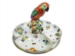 HEREND  MODELO QUEEN VICTORIA- EXUBERANTE BANDEJA PARA APERITIVOS (HORS D'OEUVRES) EM PORCELANA DA PRESTIGIADA MANUFATURA HEREND COM O HISTÓRICO E DESEJÁVEL  PADRÃO QUEEN VICTORIA  CARACTERIZADO POR RICAS COMPOSIÇÕES DE PEÔNIAS, FLORES SILVESTRES E BORBOLETAS MULTICOLORIDAS, REALÇADAS POR DELICADOS FILETES DOURADOS E RELEVOS MOLDADOS NA PORCELANA (ESTILO ROSTRATO). O CONJUNTO, HARMONIOSAMENTE ESTRTUTURADO  É COMPOSTO POR UM PRATO BASE CIRCULAR DECORADO E QUATRO TRAVESSAS REMOVÍVEIS EM FORMATO DE CRESCENTE (CRESCENT-SHAPED DISHES). O ELEMENTO CENTRAL É COROADO POR UMA EXPRESSIVA E DETALHADA ESCULTURA TRIDIMENSIONAL DE UM PAPAGAIO (ARARA) PINTADO À MÃO EM TONS VIBRANTES DE LARANJA, VERDE E AZUL, EMPOLEIRADO SOBRE UM TRONCO ESTILIZADO COM DETALHES FLORAIS. MARCA DA MANUFATURA HEREND PINTADA E IMPRESSA NA BASE DE CADA PEÇA. HUNGRIA, SEC. XX. 25,54 CM (ALTURA) POR 36,8 CM (DIAMETRO)NOTA:O padrão Victoria é um dos designs mais históricos da Herend e leva o nome da rainha homônima que se apaixonou por sua originalidade.  Originalmente, o padrão Victoria foi desenhado por Fischer Mór, fundador e proprietário da Fábrica de Porcelana Herend. O design foi introduzido em 1851, tendo raízes nos designs orientais de porcelana fina. Inicialmente, o design foi chamado de Extremo Oriente para trazer referencias da porcelana chinesa no inicio do que seria o chinoiserie europeu. A decoração é repleta de borboletas e flores estilizadas e detalhadas. As bordas são pintadas com elementos florais e acabamento em verde  ou outra cor. Durante a Grande Exposição das Obras da Indústria de Todas as Nações de 1851, no Palácio de Cristal, a primeira feira mundial, a Rainha Vitória apaixonou-se pelo design. Ela encomendou um grande conjunto de porcelana Herend, que a fábrica da Herend Hungria  na época Herend Áustria-Hungria  trabalhou durante dois anos para produzir e enviar integralmente. Gosto de brincar que a Rainha Victoria foi uma precursora dos influencers  modernos, tudo que ela gostava e usava caia nas graças do mundo. A Herend inteligentemente rebatizou o padrão Extremo Oriente para Queen Victoria o que tornou esse modelo o mais cobiçado pelos encomendantes.  A porcelana húngara da Herend logo passou a ser requisitada pela nobreza europeia para a criação de designs únicos e excepcionais. As décadas seguintes foram algumas das mais bem-sucedidas da Herend, em parte graças ao imperador austríaco Francisco José I, que concedeu a Fischer o prestigioso título de Fornecedor da Corte Real em 1865.