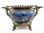 GRANDE JARDINIERE JAPONESA DO PERIODO EDO TOCUGAWA (DEC. 1860) ,   EM BRONZE COM ESMALTES CLOISONEE  ENRIQUECIDA COM MONTURE EM BRONZE ORMOLU ESTILO E ÉPOCA NAPOLEÃO III.  OS EXUBERANTES ESMALTES APRESENTAM CRISANTEMOS, PEONIAS, CEREJEIRAS E PÁSSAROS. O MONTURE TEM FIGURAS DE DRAGÕES A GUISA DE ALÇAS;. OS PÉS APRESENTAM FIGURAS DE CABEÇAS DE FERAS MÍTICAS ORIENTALISTAS.  ESTE CACHEPOT É REPLETO DE HISTÓRIA E TÉCNICAS ARTÍSTICAS FASCINANTES. M CASAMENTO ARTÍSTICO ENTRE O JAPÃO E A FRANÇA. ESTA PEÇA REPRESENTA O ÁPICE DO JAPONISME NA SEGUNDA METADE DO SÉCULO XIX, UM MOVIMENTO EM QUE A EUROPA FICOU TOTALMENTE OBCECADA PELA ARTE ASIÁTICA. O VASO CENTRAL DE ESMALTE FOI PRODUZIDO POR ARTESÃOS NO JAPÃO (DA ERA MEIJI), MAS A RICA ESTRUTURA METÁLICA EXTERNA, COM OS GRIFOS E ADORNOS RENDADOS, FOI FUNDIDA E MONTADA POR RENOMADOS PRATEIROS E BRONZISTAS NA FRANÇA. ERA COMUM QUE GRANDES FUNDIÇÕES FRANCESAS, COMO A MAISON BARBEDIENNE OU A FAMOSA LOJA L'ESCALIER DE CRISTAL, COMPRASSEM AS PEÇAS ORIENTAIS PARA CRIAR ESSAS MONTAGENS LUXUOSAS PARA A ELITE EUROPEIA. FRANÇA E JAPÃO DEC. DE 1860/70. 45 X 29 CMNOTA: NOTA: A Técnica do Cloisonné é Puro Trabalho de Paciência. O termo Cloisonné vem do francês cloison, que significa "partição" ou "divisão".O desenho dos pássaros, crisântemos e borboletas não é apenas pintado: o artesão cola delicados fios metálicos (geralmente de latão, cobre ou prata) sobre o corpo de metal da peça para formar as silhuetas. Em seguida, cada pequeno espaço criado pelos fios é preenchido manualmente com uma pasta de pó de vidro colorido (o esmalte).A peça vai ao forno a cerca de 800C, o vidro derrete e encolhe, exigindo que o processo de preenchimento e queima seja repetido várias vezes até ficar plano. Por fim, a superfície é polida exaustivamente até atingir o brilho do vidro. Na cultura oriental, os elementos retratados no esmalte azul turquesa carregam fortes simbolismos: Crisântemos brancos e flores: Representam a longevidade, a nobreza e o outono. O crisântemo é, inclusive, o símbolo da família imperial japonesa. Borboletas e Pássaros: Simbolizam a alegria, a transformação, a liberdade e a renovação da vida. As  alças e pés de bronze dourado dessas peças trazem figuras mitológicas (como dragões ou grifos estilizados). Na tradição asiática, o dragão é um ser celestial associado à proteção, à boa sorte e ao poder sobre as águas (o que faz todo sentido para um vaso feito para conter plantas e terra). Os franceses adaptaram essa estética ao gosto ocidental, adicionando asas e detalhes robustos inspirados no estilo renascentista e barroco.  O acabamento dourado do bronze (conhecido como ormolu) era feito através de uma técnica francesa clássica que misturava ouro com mercúrio. A mistura era aplicada ao bronze e depois aquecida para que o mercúrio evaporasse, deixando apenas uma camada reluzente de ouro puríssimo. Devido à extrema toxicidade dos vapores de mercúrio exalados pelos artesãos, esse método de douramento foi proibido na França ainda no século XIX, tornando as peças remanescentes autênticas relíquias de uma era artesanal extinta.