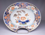 REINADO KANGXI  (1662-1722)  MUITO RARA E EXPLÊNDIDA BACIA DE BARBEAR EM PORCELANA DECORADA COM O VIBRANTE PADRÃ0 CHINA-IMARI. EXTRAORDINÁRIA PEÇA AINDA DO FINAL DO SEC. XVII. APRESENTA UM RECORTE OU REENTRÂNCIA DISTINTIVA EM FORMA DE CRESCENTE AO LONGO DA BORDA, QUE PERMITIA AO USUÁRIO SEGURAR O RECIPIENTE FIRMEMENTE CONTRA O PESCOÇO PARA COLETAR ÁGUA COM SABÃO, ESPUMA OU SANGUE DURANTE PROCEDIMENTOS DE HIGIENE PESSOAL E MÉDICOS. TEM CONCAVIDADE PRONUNCIADA PARA RETER A ÁGUA E ESPUMA DE AFEITAR MAS TAMBÉM O SANGUE TÍPICOS DO OFÍCIO DE BARBEIRO DESTE PERÍODO. A EXUBERANTE DECORAÇÃO CHINA IMARI APRESENTA A BORDA EM AZUL COM RESERVAS CONTENDO ARRANJOS FLORAIS EM ROUGE DE FEUR REMATADOS EM RICO OURO. A CALDEIRA TEM GRANDE RESERVA COM SALGUEIRO E VISTOSA PEÔNIA E OUTRAS FLORES E RAMAGENS. A PARTE TRASEIRA DA PEÇA TAMBÉM É DECORADA COM FLORES. APRESENTA DOIS FUROS CARACTERÍSTICOS PERFURADOS NA BASE SEMPRE PRESENTES NAS PEÇAS DESSE GÊNERO QUE SÃO ORIGINAIS. A FUNÇÃO DESSES FUROS É PERMITIR A PASSAGEM DE UM FIO METÁLICO PARA QUE FOSSE PRESA A PAREDE QUANDO O BARBEIRO NÃO A ESTAVA UTLIZANDO POSSUINDO DESSA FORMA UMA FUNÇÃO ESTÉTICA E DE OSTENAÇÃO. IMAGINE A RARIDADE DE UMA PEÇA DE PORCELANA CHINESA NESSA ÉPOCA PARA O OFICIO DO BARBEIRO, O PREÇO ERA CERTMAENTE VULTOSO DEMAIS PARA QUASE A TOTALIDADE DOS PROFISSIONAIS. ESTA TIGELA DE BARBEIRO É UM EXCELENTE EXEMPLO DE UM OBJETO DE USO COTIDIANO FABRICADO NA CHINA, DE ACORDO COM O GOSTO OCIDENTAL, PARA OS MERCADOS DE CLASSE ALTA DAS AMÉRICAS E DA EUROPA. COMO OUTROS PRODUTOS DE EXPORTAÇÃO ASIÁTICOS, ELA TERIA SIDO COMERCIALIZADA PELA COMPANHIA DAS INDIAS OCIDENTAIS. CHINA, FINAL DO SEC. XVII. 35 CM DE COMPRIMENTONOTA: No século XVIII, o ofício de barbeiro era uma profissão multifuncional, dramática e muito distante do conceito de bem-estar e estética que temos hoje. Eles eram conhecidos como barbeiros-cirurgiões e formavam a linha de frente do atendimento de saúde da população europeia e colonial. Os barbeiros eram responsáveis por procedimentos médicos invasivos porque os médicos acadêmicos (formados em universidades) consideravam o trabalho manual e o contato com o sangue algo inferior. Os procedimentos cirúrgicos dos barbeiros envolviam: SANGRIAS: Era o procedimento mais comum. Acreditava-se que a maioria das doenças era causada pelo excesso de sangue no corpo. O barbeiro cortava uma veia do paciente e colhia o sangue na bacia de porcelana ou latão. EXTRAÇÃO DE DENTES: Sem anestesia, os barbeiros arrancavam dentes estragados usando ferramentas brutas como a "chave de Garengeot". PEQUENAS CIRURGIAS: Tratavam de feridas de guerra, amputavam membros inflamados, tratavam fraturas e cortavam abscessos. O famoso poste listrado em vermelho, branco e azul que vemos hoje nas barbearias nasceu justamente dessa rotina médica do século XVIII:  O paciente segurava firmemente um bastão de madeira para fazer as veias do braço saltarem para a sangria.  Após o procedimento, as bandagens sujas de sangue eram lavadas e colocadas para secar, enroladas em volta desse bastão do lado de fora da loja. O vento girava as bandagens no bastão, criando o padrão listrado. O vermelho representava o sangue arterial, o azul as veias, e o branco as bandagens limpas. O século XVIII foi o período de crise e transição para a profissão, marcado por uma intensa disputa de classes:Barbeiros-Cirurgiões de elite: Atendiam a nobreza, tinham lojas sofisticadas e usavam bacias de porcelana chinesa importada para demonstrar status. Barbeiros ambulantes: Atendiam os pobres nas ruas ou mercados, carregando suas ferramentas em uma bolsa e uma bacia de latão barata sob o braço. Em meados do século XVIII (em 1745 na Inglaterra e 1743 na França), decretos reais separaram oficialmente os cirurgiões dos barbeiros. Os cirurgiões fundaram suas próprias corporações científicas, e os barbeiros perderam o direito de realizar grandes cirurgias, focando mais no cabelo, barbas e perucas.Com a perda das funções médicas no final do século, os barbeiros se reinventaram focando na moda. O século XVIII foi o auge das perucas brancas empoadas para homens e mulheres da aristocracia. O barbeiro passava horas modelando, passando pomada (feita de banha de porco) e jogando pó de arroz ou amido sobre as perucas dos clientes.