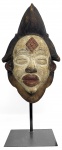 MASCARA PUNO - GABÃO -  TRADICIONALMENTE PATINADAS EM BRANCO COM CAULIM E CINGINDO ELABORABOS PENTEADOS ESSAS MÁSCARAS CARREGAM UM SIGNIFICADO ESPIRITUAL PROFUNDO PARA O POVO PUNO  GABÃO, INICIO DO SEC. XX. 32 CM DE ALTURA (SEM CONSIDERAR A ALTURA DA BASE QUE ACOMPANHA A PEÇA)NOTA: O povo Punu do Gabão e da República do Congo vive em pequenas aldeias, principalmente ao longo da bacia do rio Ogowe. São um povo matrilineal organizado em chefaturas hereditárias. Como a maioria dos povos da África central e noroeste, possuem crenças religiosas animistas. Em cada aldeia Punu existe a Sociedade Mwiri, uma organização secreta masculina cujos únicos membros podem confeccionar máscaras. As máscaras desempenham muitas funções importantes na sociedade Punu, incluindo o controle social e a purificação de espíritos malignos e feitiçaria. A máscara mukudj (também chamada okuyi , mokoi , ukuyi , mokoi ou mbwanda ) representa simultaneamente um espírito ancestral e uma mulher idealizada. O padrão de losangos na testa e os quadrados nas têmporas representam as marcas de escarificação comuns entre as mulheres Punu e enfatizam a simetria do rosto. O mukudj é dançado por membros masculinos da Sociedade Mwiri em pernas de pau muito altas em nascimentos, funerais, cerimônias de iniciação à vida adulta e outros eventos sociais importantes para invocar a aprovação dos espíritos ancestrais.