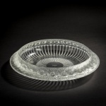 LALIQUE  PADRÃO MARGUERITES (MODELO Nº 10-403).  EXPLÊNDIDO BOWL EM CRISTAL DA MANUFATURA DE LALIQUE (ASSINADA SOB A BASE).  PROJETADA ORIGINALMENTE COMO UM CENTRO DE MESA PARA COMPOSIÇÕES FLORAIS, FRUTAS OU COMO PEÇA DECORATIVA AUTÔNOMA PARA APARADORES E MESAS DE JANTAR REFINADAS. A BORDA EXTERNA APRESENTA UMA ESTRUTURA CONTÍNUA E DENSAMENTE MOLDADA COM FLORES DE MARGARIDA FOSCAS EXIBINDO DETALHES INTRINCADOS NAS PÉTALAS E CENTROS TEXTURIZADOS. BAIXO DA BORDA, A TAÇA TRANSITA PARA UM VIDRO TRANSPARENTE COM RANHURAS LINEARES RADIANTES ( GODRONS ) QUE DIRECIONAM A LUZ PARA O CENTRO. UM TESTEMUNHO DA MAESTRIA DE LALIQUE QUE CONTRASTA O RELEVO FOSCO COM ACABAMENTO ACETINADO AO VIDRO TRANSPARENTE E POLIDO PARA CRIAR PROFUNDIDADE E BRINCAR COM A LUZ. AS MARGARIDAS SÃO UM TEMA MUITO QUERIDO POR RENÉ LALIQUE. ELE DESENHOU ESTA OBRA PELA PRIMEIRA VEZ EM 1933. ESTA PEÇA DE MEADOS DO SEC. XX ESTÁ EM EXCELENTE CONDIÇÃO! FRANÇA, MEADOS DO SEC. XX. 36 CM DE DIAMETRO