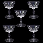 BACCARAT POLIGNAC   COM SELO DA MANUFATURA  .IDENTICAS AS OFERTADAS NO LOTE ANTERIOR. ORIGINALMENTE ENCOMENDA DO GOVERNO FRANCÊS (PRESIDENTE RENÉ COTY) PARA O BANQUETE OFICIAL OFERECIDO EM HOMENAGEM A RAINHA ELIZABETH II NA GALERIA DOS ESPLEHOS DO PALÁCIO DE VERSALHES EM 9 DE ABRIL DE 1957. RÉGIO CONJUNTO COM CINCO TAÇAS PARA CHAMPAGNE EM CRISTAL DO MODELO POLIGNAC DA BACCARAT. APRESENTA LAPIDAÇÕES VERTICAIS ESPESSAS E PROFUNDAS (ORGUE BEVEL CUTS) QUE SAEM DA HASTE E SOBEM PELO BOJO. O DETALHAMENTO GEOMÉTRICO CRIA UM VISUAL ROBUSTO QUE SIMULA UM CÁLICE MODERNO, CONTRASTANDO COM A SUAVIDADE DA BORDA SUPERIOR LISA. FOI BATIZADO EM HOMENAGEM À DUQUESA DE POLIGNAC, FIGURA HISTÓRICA CONHECIDA POR SEU GOSTO REFINADO E PROXIMIDADE COM A RAINHA MARIA ANTONIETA. FRANÇA, DEC. 1950. 12 X 10,5 CMNOTA: O modelo foi criado exclusivamente para servir a Rainha Elizabeth II e o Príncipe Philip em sua visita oficial à França em 1957. Ele decorou a mesa do banquete na Galeria dos Espelhos no Palácio de Versalhes. O presidente francês René Coty encomendou o design para impressionar a monarca com a modernidade do cristal francês. Seus cortes verticais profundos funcionam como prismas que refletem a luz das velas, criando um brilho único na mesa. Cada taça exigia o trabalho de vários artesãos altamente qualificados, conhecidos como Meilleurs Ouvriers de France.do: Devido à lapidação profunda, as taças são notavelmente mais pesadas que os modelos tradicionais. O nome celebra a Duquesa de Polignac, que foi a amiga mais íntima e favorita da rainha Maria Antonieta. As peças produzidas antes de 1936 não tinham logotipo gravado, mas como o Polignac nasceu nos anos 1950, todas as peças originais trazem a marca circular jateada da Baccarat na base.