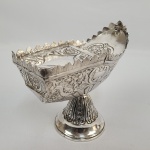 FABULOSA NAVETA DE GRANDE DIMENSÃO EM PRATA DE LEI DA BAHIA. REQUINTADO TRABALHO SETECENTISTA. COM DESEJÁVEL FEITIO DE NAVE É DECORADA AO ESTILO DOM JOÃO V COM ROCAILLES E FLORES. "O CONVÉS" TEM TAMPA BASCULANTE LISA COM BELISSIMA RESERVA CENTRAL COM FLORES E RAMAGENS. A POPA TEM ESCULELA LISA CERCADA POR ROCAILLES VEGETALISTAS. TODA A PARTE SUPERIOR TEM CERCADURA EM RECOERTADA SIMULANDO EXTREMIDADES DE FOLHAS. BASE CAMPANULAR REMATADA POR FOLHAS DE ACANTO NO SENTIDO VERTICAL.. BAHIA, SEC. XVIII. 18 X 19 CM