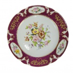 DOM PEDRO I - SERVIÇO DA BARRA BORDEAUX - PRATO EM PORCELANA  FRANCESA EM PASTA DURA, PERTENCENTE AO SERVIÇO  DE D. PEDRO I. PASTA DURA. ABA DECORADO COM ESMALTES BORDO QUE NOMEIAM O SERVIÇO DECORADOS COM RESERVAS CONTENDO FLORES EM ESMALTES MANUAIS  ENTREMEADAS POR ROCAILLES EM OURO. A CALDEIRA TEM EXUBERANTE CORBÉLIA FLORA. PRINCIPIO DO SÉC. XIX. EXEMPLARES DESSA LOUÇA COMPÕE O ACERVO DO PALÁCIO IMPERIAL DE PETRÓPOLIS, MUSEU HISTÓRICO NACIOONAL. MAIS DE CEM PEÇAS DESSE SERVIÇO COMPÕE O ACERVO DA FUNDAÇÃO MARIA LUIZA E OSCAR AMERICANO. FINALMENTE, . PEÇA DO MESMO SERVIÇO ESTÁ REPRODUZIDA NA PÁGINA 190 DO LIVRO LOUÇA DA ARISTOCRACIA NO BRASIL, DE JENNY DREYFUS. 23 CM DE DIAMETRO. NOTA: O ANUÁRIO DO MUSEU IMPERIAL DE PETRÓPOLIS DE 1942 TRAZ REFERÊNCIAS AS PORCELANAS BRAGANTINAS E IMPERIAS E SEUS REGISTROS ALÉM DE REMEMORAR OS DRAMÁTICOS LANCES DOS LEILÕES DOS PAÇOS IMPERIAS OCORRIDOS LOGO APÓS O NEFASTO GOLPE REPUBLICANO : Foram muitos, e incontestavelmente, os aparelhos de porcelana da Casa de Bragança. Houve-os de todas as formas e pinturas, das mais variadas marcas. Ricos e pobres. Suntuosos e singelos. Desde os mais preciosos espécimes originários da remota China, trazidos por d. João VI (os serviços dos galos, do pastor, das corças e dos pavões), além de uma multiplicidade de outras peças de antigo cunho europeu, onde avultavam decorações de palácios, quintas e castelos, pássaros e quadrúpedes em combinações harmoniosas. São muito de destacar, em serviços do Primeiro Reinado, os que trazem a marca da duquesa dAngoulême, essa fidalga flor-delis de França, filha de Luiz XVI, enérgica e impulsiva, de quem Napoleão dizia ser o único homem da família. Foram muitos. Das mais soberbas e preconizadas marcas. Sèvres, Saxe, Bourg-la-Reine, Chantilly, Sarreguemines, Capo di Monte, o vieux Limoges. Esses que passaram mais tarde das mãos reais à tradição da família brasileira, especialmente daquelas castas mais achegadas ao paço, das saudosas matronas patrícias que foram amigas íntimas da princesa Isabel e d. Teresa Cristina, e no leilão do Paço Imperial atropelavam o paciente Virgílio  com um chorado peditório de um sapato velho, que a medo escondiam no seio, um lenço usado, um grampo de chapéu, uma negalha de cabelo ainda agarrado à tartaruga dos pentes, qualquer coisinha, um pequeno nada que tivesse pertencido à mãe dos brasileiros.  Ouvir o amigo Virgílio, que ainda está vivo e são, com a memória bem fresca, no relato de episódios a que assistiu na sua mocidade, no acidentado e trabalhoso campo do seu árduo mister, é o mesmo que folhear um livro de impressões, de comum decoradas com tintas vivas, as próprias tintas de que é tão rica sua paleta de pintor. Foi ele, ao que se viu, um dos martelos desse leilão insigne, quem primeiro lançou aos ares da cidade, entre as árvores robustas da velha quinta imperial, o pregão melancólico das grandes coisas históricas, o desbarato inicial do regime que se impunha.  Quanto dão por isto? E por aquilo?  gritava o arauto, erguendo sobre as cabeças atônitas os mais caros e prezados ornamentos do Brasil monárquico. E os candidatos acorriam, nervosos, febricitantes. Disputavam as peças propaladas com esse afã tumultuário de casa de jogo que tem todos os leilões. Muitos, aterrorizados, lançavam medrosamente as ofertas ou as mandavam lançar  não fosse estar por ali, detrás daqueles reposteiros armoriados, algum cérbero republicano. Outros, jocosos, em geral aderentes de última fornada, como emplastros caseiros que se agarram a qualquer protuberância, faziam troça, metiam a ridículo a coisa apregoada, achincalhavam o palácio, o imperador, os príncipes, a monarquia, sem saber, na sua alta ignorância, que zombavam da própria história pátria. Virgílio, braço direito do velho J. Dias, ia cumprindo, indiferente, o seu dever. Agora, um objeto de sensação. Um piano de cauda, todo em jacarandá, com esculturas e filetes dourados, da melhor fábrica inglesa. Quanto vale, senhores? Foi da princesa Isabel. Nele tocou o grande Gottschalk. E para garantir as vozes do instrumento, ele mesmo em pessoa larga o martelo inseparável, assenta-se à banqueta estofada e executa com graça e petulância um trecho do seu repertório. Sucesso! Os assistentes aplaudem. Abraçam o concertista improvisado. E o piano famoso alcança a soma exorbitante de dois contos de réis. Por fim, o leilão termina. Durara vários dias. Fora o assunto de todas as conversas, no Parlamento, nos cafés, nos clubes, nos lares, nas ruas da cidade. Fiel ao trono deposto, a velha-guarda, que comparecia diariamente àqueles desoladores espetáculos, comprava o que podia, de rosto grave, compungido, ufana de ainda poder acautelar alguma coisa que houvesse sido honrada com o carinho paternal de Suas Majestades. Por fim, todo mundo dispersa. Vai-se tornando deserto o parque antigo de Elias Lopes. Deserto o casarão arquejante de tanta passada inútil pelos seus salões dourados, pelos seus longos e sombrios corredores. Deserto o jardim bonançoso onde os pássaros chilreiam, sob as ramadas hospitaleiras, por sobre as águas livres e cantantes. Daqui, dali, montando guarda aos destroços da monarquia, um ou outro soldado, a sentinela sonolenta daqueles pousos abandonados. Ao cabo do dramático episódio, já cerrando as portas, Virgílio, certa manhã dessa tristonha primavera, desce ao fundo do paço, pára em frente de um tapume de madeira, desses escuros vãos de escada onde os jardineiros costumam recolher as ferramentas. Espia por uma fresta. Vê lá dentro alguma coisa que brilha. Força a entrada, e dá com os olhos em quatro enormes caixas de Flandres. Consulta as suas notas. Corre os lotes desprezados por sua insignificância entre panelas velhas, cacarecos e trastes imprestáveis. Lá está: quatro latas de folha, avaliadas a seiscentos réis. O lote inteiro, dois mil e quatrocentos. Mas afinal, que será? Só isso teriam dado pelas caixas. O fato é que elas estavam pesadas. Conteriam alguma coisa útil, quem sabe, alguma coisa de valor? Assim fechadas, lacradas com os sinetes do Império... Não. Vamos a ver que diabo se intromete nelas? E abre uma caixa: salta um berço de criança. E que berço, santo Deus! Que maravilha de arte, com as suas esculturas de pau santo, os seus anjinhos esvoaçantes, as suas bolas de marfim legítimo! Que se abra a outra lata. Outro berço. Igualzinho ao primeiro, com o mesmo luxo, o mesmo estilo, a mesma graciosidade. E assim o terceiro e o quarto. Aturdido, Virgílio chama um velho servidor do paço, que por ali ainda se detinha, a capinar distraidamente, mero pretexto de matar saudades.  Que é isto, meu amigo?  Isto, seu moço? O sinhô antão num sabe o que é isso? São os berços dos príncipes! Muito esta mão véia cansada embalô eles... E eram mesmo. Os bercinhos gentis dos herdeiros do trono. Ali jogados como inutilidades, os casulos de ouro e seda que tanto sonho adormentaram
