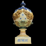 FÁBRICA DE DEVESAS JOSÉ P. VALENTE -  VILA NO DE GAIA  PORTO - MONUMENTAL PINHA ESTATUÁRIA EM FAIANÇA DE DEVESAS. FUNDO ESMALTADO É DECORADA A TODA VOLTA COM GUIRLANDA RELEVADA EM AMARELO E AZUL. TAMBÉM FESTÕES E ESTRELAS. BASE TEM INSCRIÇÕES EM AZUL DA FÁBRICA DE DEVESAS E DO FABRICANTE JOSÉ P VALENTE. CONSTRUÍDAS COMO PEÇAS DE APARATO, SEGUNDO A TRADIÇÃO DECORATIVA PORTUGUESA E EUROPEIA, A PINHA SIMBOLIZA A PROSPERIDADE, A IMORTALIDADE, A FORÇA VITAL E A BOAS-VINDAS.. PORTUGAL,SEC. XIX. 84 CM DE ALTURANOTA:  A Fábrica das Devesas, fundada em 1865 em Vila Nova de Gaia, tornou-se uma das maiores e mais importantes fábricas de cerâmica de toda a Península Ibérica. Além das pinhas, a fábrica era famosa por produzir estátuas gigantes, fontes de jardim, fontanários e decorações de fachadas que ainda hoje adornam ruas em Portugal e no Brasil. A empresa publicava catálogos ilustrados impressionantes para a época, permitindo que clientes de colónias e outros países encomendassem peças de arte por catálogo. Na arquitetura tradicional portuguesa, colocar pinhas de cerâmica nos portões de entrada, muros ou no topo das casas era um símbolo universal de hospitalidade e acolhimento. Desde a antiguidade (associada ao deus Baco), a pinha representa a fertilidade, a regeneração, a imortalidade e a prosperidade devido à sua abundância de sementes (pinhões). No Brasil, devido ao formato esculpido e à distância da Europa, muitas destas pinhas importadas no século XIX e início do XX passaram a ser chamadas popularmente de "pinhas-de-açúcar" ou até confundidas visualmente com representações do ananás (abacaxi), que também simboliza hospitalidade. 