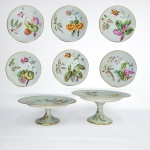 PORCELANA DO PERÍODO GEORGE III -  MARCAS INCISAS ATRIBUIDAS A PORCLEANA DE CAUGHLEY (S INCISO REPRESENTANDO A PALAVRA SALOPIAM) E NUMERAÇÃO APLICADA MANUALMENTE. EXUBERANTE SERVIÇO PARA SOBREMESA EM PORCELANA COM DECORAÇÃO BOTÂNICA SOB FUNDO CELADON REMATADO EM OURO. APRESENTA FLORES EXOTICAS E SAMAMBAIS . COMPOSTO POR OITO PEÇAS: SÃO DOIS SUPORTES PARA SERVIÇO (COMPORTS) COM PÉS ELEVADOS EM FEITIO DE TAÇA INVERTIDA. TAMBEM SEIS PRATOS. A DECORAÇÃO É EXPLENDIDA, ELEGANTE E APAIXONANTE. INGLATERRA, FINAL DO SEC. XVIII. OS PRATOS TEM 23 CM DE DIAMETRO E OS COMPORTS TEM 23 X 12 CM. NOTA: As louças com decoração botânica no período georgiano na Inglaterra refletem o interesse científico e artístico da época pelas plantas, destacando-se por pinturas manuais detalhadas, uso da técnica de transferência (transferware) e inspiração em enciclopédias da natureza. A fascinação por botânica e jardinagem impulsionou a reprodução fiel de flores, folhas e espécimes reais nas peças de cerâmica e porcelana. As Fábricas usavam a pintura manual realista quanto a impressão por transferência em cobre, permitindo traços finos em massa branca. A porcelana com esmaltes simulando celadon aperfeiçoada no final do período, trazia uma base clara e translúcida que valorizava as cores das plantas.Principais Manufaturas: Chelsea e Derby: Pioneiras na pintura floral delicada e naturalista inspirada no estilo Rococó inicial. Worcester: Conhecida por buquês botânicos coloridos e detalhes dourados sobre fundos esmaltados. Wedgwood: Fundada por Josiah Wedgwood, inovou na cerâmica refinada com relevos e motivos inspirados na natureza e na antiguidade