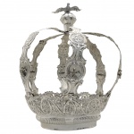 IMPÉRIO DO DIVINO ESPÍRITO SANTO - MAGNÍFICA COROA EM PRATA DE LEI DECORADA COM FLORES E RAMAGENS DISPOSTAS ENTRE ELEGANTES ROCAILLES.. CINCO HASTES REUNIDAS  SOB ORBE COROADA COM DIVINO ESPÍRITO SANTO EM REALÍSTICO FEITIO DE PÁSSARO COM ASAS ABERTAS. BASE FENESTRADA ESTILO DONA MARIA I. BRASIL, SEC. XIX.   26 CM DE ALTURA. 360 GNOTA: Este é um fato pitoresco ocorrido na Pindamonhangaba do início do século XIX. O pano de fundo do acontecimento é a Festa do Divino Espírito Santo. Em 4 de agosto de 1827 foi lavrado na VILLA DE NOSSA SENHORA DO BOM SUCESSO DE PINDAMONHANGABA um relato Nossa fonte é um documento datado de 4 de agosto de 1827, os autos de um processo legal. Naquele ano de 1827, o cidadão Salvador Francisco de Tolledo foi a juízo com a intenção de receber a quantia de oito mil réis que o padre Luís Justino Velho Columbreiro lhe devia e morrera sem lhe pagar. O padre mencionado, na ocasião em que contraíra a dívida era vigário colado (pároco) da Vila Real de Nossa Senhora do Bom Sucesso de Pindamonhangaba, mas foi na qualidade de imperador da Festa do Divino que havia acertado com Salvador de Tolledo que lhe pagaria dezesseis mil réis para que o mesmo saísse pela região levando a bandeira do divino, cantando na folia e recolhendo donativos dos fiéis para a realização dos festejos ao Divino Espírito Santo. Nesta missão Toledo teria passado três meses longe de casa, andando por caminhos agros (terra cultivada ou cultivável), espinhosos e cerros (colinas penhascosas).Como pagamento pelas andanças à cata de recursos para a festa, o padre pagara-lhe somente a metade, ou seja, oito mil réis, e não fora em dinheiro, mas em fazenda na logem de José Camillo Lelles. Tomando o termo logem como casa de comércio e atentando para o complemento logem de fazenda ceca utilizado pelo escrivão na ocasião do depoimento de José Camillo Lellles na qualidade de testemunha de Tolledo, e considerando o termo fazenda como mercadorias, pode-se concluir que o estabelecimento onde o padre havia autorizado seu credor a receber em mercadorias, comercializava gêneros sólidos e não molhados como vinho azeite e outras substâncias líquidas. E fora o que o contratado pelo então imperador do divino recebera, conforme confirmação do comerciante Lelles, acrescentando que sabia que o padre ficara devendo outra parte por ser da casa do falecido vigário, no sentido de ser conhecido da família, frequentar sua residência. Além do mercador Lelles, no processo foram inquiridas como testemunhas por parte de Francisco de Tolledo, o padre Claro Vieira Cortes. Este religioso declarou estar ciente da dívida do vigário, mas não se lembrava quanto ao tempo de duração da empreitada de Tolledo, se haviam sido mesmo três meses.Outra testemunha apresentada, Joaquim Ferreira da Silva, fora um dos integrantes da comitiva do divino liderada por Tolledo, um dos companheiros de folia. Segundo este, andaram mais de três meses pelos distritos da Vila de Nossa Senhora do Bom Sucesso, Taubaté São Luís, Cunha e outras localidades na mesma diligência de pedir esmolas Joaquim não só confirmou a dívida do padre, mas afirmou ser o vigário homem muito pouco inclinado a pagar o que devia. Consta na conclusão do processo, que não tendo sido apresentada nenhuma oposição por parte de José Cerqueira César, testamenteiro do padre Justino Velho Columbreiro, o juiz Antonio Melitão de Souza Aimbere determinou que apensa aos autos de inventário do finado devedor se lhe separassem bens para seu devido pagamento, sem prejuízo de terceiro. O vigário Luís Columbreiro foi ordenado padre pelo Seminário São José, do Rio de Janeiro, em 1779 fora responsável pela paróquia de Pindamonhangaba de 1793 a 1806. Seu falecimento se dera em 25 de agosto de 1826. Levando em conta esses dados, a cobrança vinha a juízo um ano após a morte do devedor. Ainda com bases no mesmo autor, mais curioso é o fato de a dívida haver sido contraída no período em que o padre exercia a função de pároco, isto é, pelo menos 20 anos antes. Supõe-se então que o credor tenha sido cobrado ao longo de duas décadas e nada do seu vigário soltar-lhe os cobres