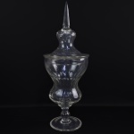 BACCARAT  GRANDE FRASCO  DE BOTICA EM CRISTAL DA MANUFATURA DE BACCARAT UTLIZADOS PARA ARMAZENAR ÁGUAS FARMACÊUTICAS. DE ELEGANTE FEITIO É LAPIDADO EM SULCOS VERTICAIS JUSTAPOSTOS. A TAMPA TEM PEGA COM FEITIO DE OBELISCO. UMA VERDADEIRA OBRA DE ARTE!  NO CONTEXTO DA FARMACOLOGIA DAS BOTICAS DO SEC. XIX. DESEMPENHAVA TAMBÉM UMA FUNÇÃO ESTÉTICA E DE STATUS FICANDO EXIBIDOS DE FORMA VISTOSA NOS SUNTUOSOS ARMÁRIOS DO ESTABELECIMENTO. O vidro incolor e límpido de alta qualidade permitia que o boticário ou o cliente conferissem rapidamente o aspecto, a cor e o nível do composto guardado. Formavam parte da cenografia de luxo das chamadas pharmacias do Segundo Reinado, atestando a sofisticação europeia do local. Valorizavam o ambiente de atendimento, transmitindo uma imagem de ciência higiênica, modernidade e confiabilidade científica aos olhos da elite que podia pagar pelos remédios importados  ELES ERAM PREENCHIDOS COM ÁGUAS COLORIDAS ARTIFICIALMENTE (EM TONS VIBRANTES DE VERMELHO, AZUL OU VERDE) E COLOCADOS NAS VITRINES. FUNCIONAVAM COMO O "LETREIRO LUMINOSO" DA ÉPOCA. QUEM PASSAVA NA RUA E VIA O GRANDE FRASCO DE CRISTAL ILUMINADO SABIA INSTANTANEAMENTE QUE ALI FUNCIONAVA UMA FARMÁCIA. OSTENTAR UMA PEÇA DESSE TAMANHO, ENCOMENDADA DA SOFISTICADA MANUFATURA FRANCESA DE BACCARAT, ERA UMA DEMONSTRAÇÃO DE IMENSO PODER FINANCEIRO DO BOTICÁRIO. OS CLIENTES DO SEGUNDO REINADO ASSOCIAVAM O BRILHO E A PRECISÃO DA LAPIDAÇÃO DO CRISTAL À PUREZA, À HIGIENE E AO RIGOR CIENTÍFICO DOS MEDICAMENTOS MANIPULADOS ALI DENTRO. FRANÇA, SEC.  XIX. 62 CM DE ALTURA (APRESENTA UM BICADO NA BORDA INTERNA DO FRASCO)
