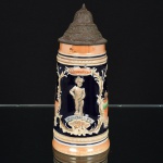 PORCELANA, um (1) tankard com ornamentação relevada e policromada sobre fundo azul cobalto, representando cena do gênero de taberna e legendas: Auch Die Freud Hat Ihre Zeit (Até Freud tem seu momento), com dispositivo musical funcionando com a música: River Kwai Marseh (Pântano do Rio Kwai), tampa em pewter, medindo 28,5 cm altura, sinais de uso, vide foto, ideal para coleção de bar.