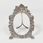FOTOGRAFIA, um (1) antigo porta-retratos oval, confeccionado em metal prateado, ornamentação relevada em concha, volutas e elementos vegetalistas, terminações em garras, faltam o vidro e suporte traseiro, possui haste expositora, sem marca de manufatura aparente, medindo 13,5 x 9,5 cm, sinais de uso, vide foto.