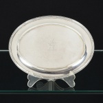 METAL PRATEADO, uma (1) bilheteira oval, ornamentação relevada no padrão Império Napoleônico com frisos intercalados por X, parte central burilada com cabeça de animal, punção não identificado na borda representando papagaio pousado em galho e a letra S, medindo 19 x 14,5 cm, sinais de uso, vide foto.