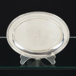 METAL PRATEADO, uma (1) bilheteira oval, ornamentação relevada no padrão Império Napoleônico com frisos intercalados por X, parte central burilada com cabeça de animal, punção não identificado na borda representando papagaio pousado em galho e a letra S, medindo 19 x 14,5 cm, sinais de uso, vide foto.