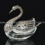 CRISTAL E PRATA INGLESA, um (1) saleiro incolor e translúcido, facetado, fundo estrelado, formato de cisne com pescoço e asas em prata, contrastada 925 milésimos, acompanha colher em prata, contrastada, com extremidade relevada em anjo e flor, medindo o saleiro 7 cm comprimento, a colher 5,5 cm comprimento, sinais de uso, vide foto.