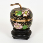 CLOISONNÉ, uma (1) caixa miniatura no formato de maça, confeccionada em metal, esmaltação policromada em elementos vegetalistas sobre fundo preto, base em madeira torneada e ebanizada, altura total 4 cm, vide foto.