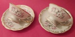 Par de delicadas xícaras de chá em porcelana Schmidt com arabescos e filetes dourados e pintura de flores e pega dourada medindo 11x7 e pires 15 cm.