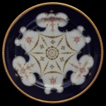 Prato em porcelana francesa da manufatura de Limoges, caracterizado por sua rica ornamentação no estilo europeu clássico, combinando um fundo em azul cobalto profundo com detalhes ornamentais em ouro dourado e detalhes florais. O miolo traz um medalhão circular cercado por um padrão radiante em formato de estrela ricamente trabalhado em ouro. Marca da manufatura no fundo. 22 cm.