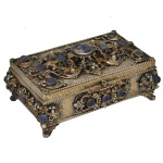 Caixa para joias de formato retangular em bronze. Apresenta frisos perolados, e ornamentação realçada por ricas composições florais com volutas, folhagens, cabochões em lápis lazúli e pérolas. Tampa seguindo o mesmo padrão com cantoneiras, encimada por pega com elegante arranjo. Quinas sustentadas por pequenos pés recurvos. Apresenta numeração sob um dos pés.