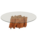 Mesa de centro rústica com base de tronco de árvore natural e tampo redondo de vidro. Esse estilo de mobiliário valoriza o design orgânico, onde o formato bruto e as texturas da madeira maciça servem como escultura central da sala. O uso do vidro temperado transparente permite total visibilidade dos detalhes e das curvas da madeira vista de cima, trazendo leveza visual ao ambiente.