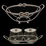 Lote constando de 2 peças, sendo: A) Par de açucareiros de vidro com base em metal prateado.  Formato navicular.10 x 25 x 10 cm. B) Suporte para travessa em metal prateado. Projetado para segurar travessas de servir quentes ou frias. Estrutura com alças e detalhes decorativos, como nós, feitos para adicionar elegância à mesa.10 x 25 x 22 cm