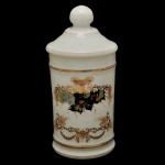 Pote de farmácia em porcelana, de formato cilíndrico. Fundo de superfícies branca com frisos dourados e decoração central com guirlandas florais em policromia. Tampa com puxador esférico. 26 x 13 cm.