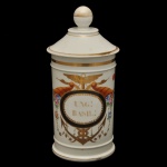 Pote de farmácia em porcelana, corpo cilíndrico com superficie branca com decorações em esmalte preto, dourado e vermelho (rouge de fer), incluindo uma tampa abobadada com frisos em dourado.  A inscrição no rotulo UNG.: BASIL moldurados com elementos decorativos como folhas de acanto. 26 x 13 cm.