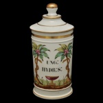 Pote de farmácia em porcelana, de forma cilíndrica. Apresenta da inscrição UNG: HYDR:S: envolto em decoração de palmeiras ladeadas por taça e serpente, símbolos clássicos da farmácia. Tampa com frisos e detalhes em ouro na borda. 26 x 12 cm.