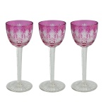 Conjunto de 3 copos de pé alto em cristal. Bojos no formato de tulipas, ornados em overlay de cor magenta, lapidados e ornamentados por treliças e raiados distribuídos em faixas verticais. Hastes retas, facetadas e serrilhadas, apoiadas em bases circulares lisas realçadas por finos gomos. 20 cm.