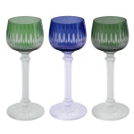 Conjunto de 3 copos de pé alto em cristal. Bojos no formato de tulipas, ornados em overlay, 2 de cor verde e o outro em azul, lapidados e ornamentados por adornos geométricos alongados sobre frisos estilizados. Hastes retas e facetadas, rematadas por anelados, e apoiadas em bases circulares realçadas por finos gomos. Um deles com bicado no pé. 19 cm.