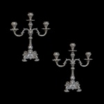 Par de candelabros em prata de lei portuguesa, contrastada, para três lumes (ou braços). Haste central decorada que sustenta três lumes para velas dispostos de forma simétrica. Apresenta rica ornamentação com elementos em relevo, ornamentos florais e volutas na base de apoio, remetendo ao estilo barroco ou vitoriano. 35 x 30 x 14 cm. Peso total 3k600 gramas.