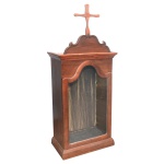 Oratório de mesa de formato retangular em madeira lustrada e lavrada. Frente com porta envidraçada com topo em arco e encimada por largo friso em degraus. Frontão recortado centralizando cruz estilizada. Base lisa e reta. Brasil. Séc. XX. 120 x 50 x 26 cm.
