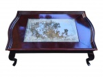 MOBILIÁRIO, uma (1) mesa de centro confeccionada em madeira escura, tampo retangular com centro em vidro translucido com gravação floral em tom fosco, apresentando bicado no vidro, pés curvos e saia reta, medindo 35x120x90cm.