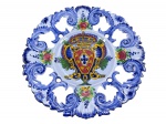 VESTAL (PORTUGAL, fundada em 1947 na Vestiaria), um (1) prato decorativo confeccionado em faiança estilo Alcobaça vitrificada na tonalidade azul, decorado por brasão, elementos florais e vegetalistas em policromia com recortes vazados, marcado no verso, medindo 29cm.