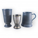 ESTANHOS e METAL PRATEADO, lote com três (3) peças, sendo: A) 1 caneca confeccionada em estanho, leves desgastes, medindo 11,5x11x7,5cm, B) 1 taça para vinho confeccionada em estanho, apresentando mossa na base, vide foto, medindo 14x7,5cm e C) 1 taça para vinho confeccionada em metal espessurado a prata com falhas na prateação, haste cinzelada por elementos florais em vazados, oxidação interna, medindo 10x6cm.