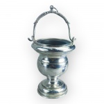 ARTE SACRA, uma (1) caldeira para água benta confeccionada em metal espessurado a prata com corpo torneado, alça central articulável, cinzelada por folhagens, apresenta falhas na prateação, recipiente interno removível, ausência do espargidor, medindo 26x13x10,5cm.